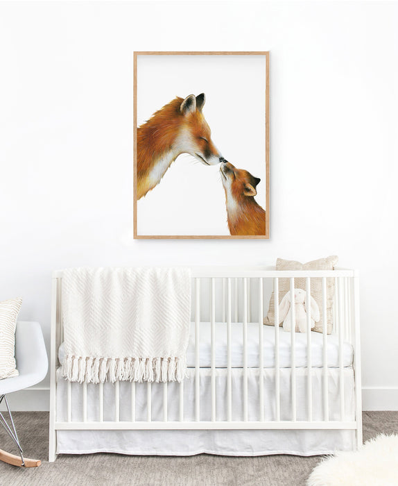 A drawing of a mama fox and her cub touching noses on a white background in a teak frame hanging in a nursery above a cot